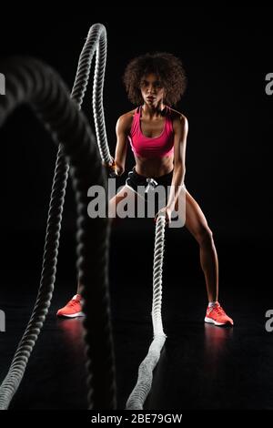 Immagine di atletica sportiva afro-americana facendo esercizio con corde di battaglia isolate su sfondo nero Foto Stock