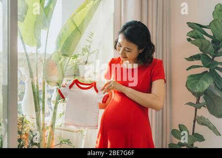 Giovane donna incinta in piedi vicino alla grande finestra con i vestiti del suo bambino futuro Foto Stock