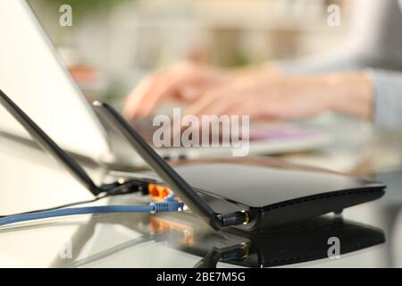 Primo piano del router a banda larga wifi che fornisce la connessione alle mani della donna utilizzando il laptop su una scrivania a casa Foto Stock