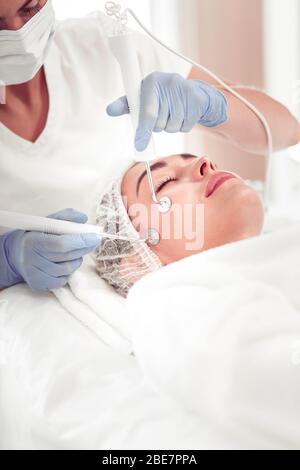 Cosmetologo in uniforme. Cosmetologa che indossa uniforme bianca che fa pulizia del viso per la donna che chiude gli occhi Foto Stock