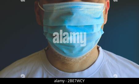 Uomo di mezza faccia che indossa la maschera blu medica durante il blocco epidemico del virus che posa sulla parete blu. Foto Stock