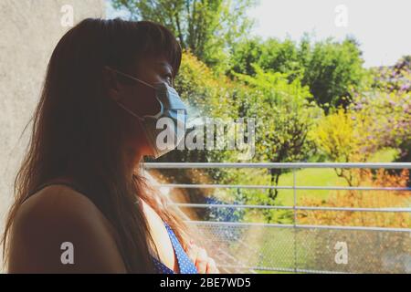 Donna asiatica annoiato che indossa maschera viso guardando fuori finestra durante la quarantena a casa Foto Stock