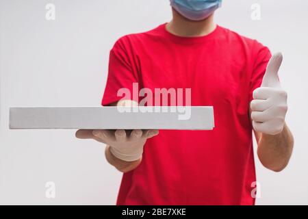 Uomo dal servizio di consegna in t-shirt rossa, indossando maschera protettiva e guanti che tengono la scatola della pizza Foto Stock