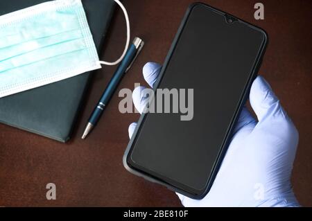 Mano in guanto monouso che tiene il telefono mobile sopra una scrivania-business durante il concetto di pandemics Foto Stock