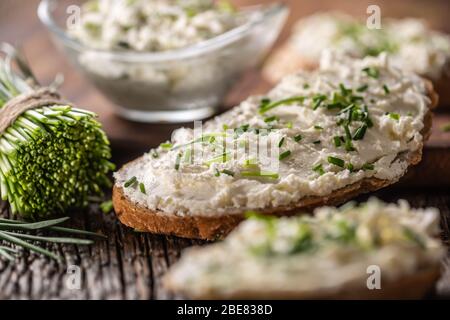 Formaggio cremoso di erba cipollina spalmato su una fetta di pane accanto a un mazzo di erba cipollina tagliata di fresco e una ciotola di spargimento sullo sfondo. Foto Stock