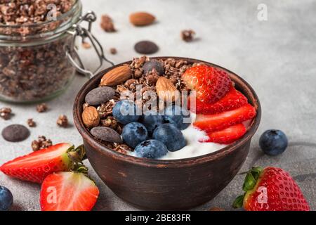 granola al cioccolato con yogurt greco, frutti di bosco freschi e noci in una ciotola su sfondo grigio per una colazione sana. primo piano Foto Stock