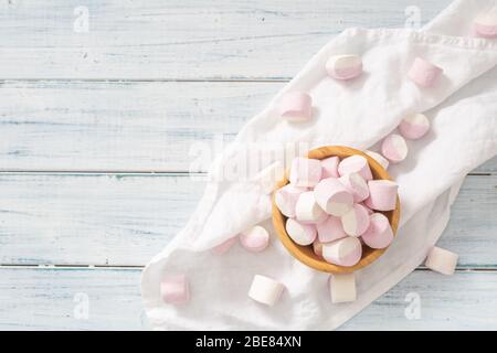 Vista dall'alto di una ciotola di legno piena di marshmallow rosa e bianco con alcuni sparsi intorno su un panno da tavolo bianco e superficie di legno bianco. Foto Stock