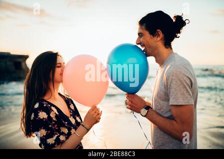 Coppia in piedi data all'aperto in spiaggia con mongolfiera. Giovane uomo e donna con palloncini sorridenti in spiaggia. Foto Stock