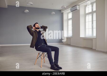 Giovane imprenditore maschile attento in abiti eleganti sognando mentre si siede su sedia in una nuova e moderna sala ufficio vuota Foto Stock