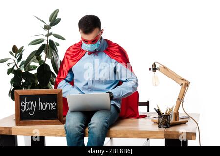 freelancer in maschera medica e costume supereroe utilizzando il laptop vicino a bordo di gesso con stay home lettere isolato su bianco Foto Stock