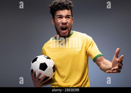 tifoso di football afroamericano arrabbiato in t-shirt gialla urlando e tenendo palla su grigio Foto Stock