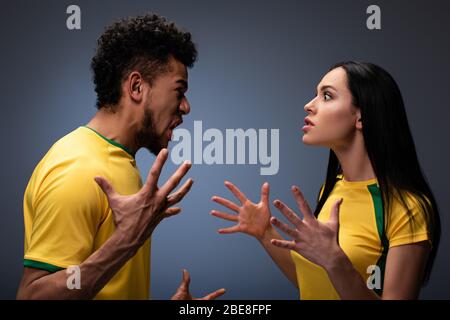 coppia aggressiva multiculturale di tifosi di calcio in giallo t-shirt gesturing e gridando su grigio Foto Stock