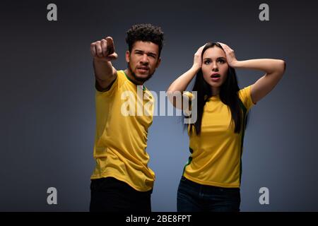 Coppia multietnica preoccupata di tifosi di calcio in magliette gialle puntando sul grigio Foto Stock