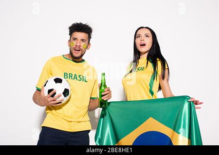 coppia multiculturale di appassionati di calcio sorpresi con bandiera brasiliana, palla e bottiglia di birra sul grigio Foto Stock