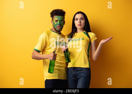 maschio african american football fan con faccia dipinta e confusa ragazza che tiene bottiglie di birra su giallo Foto Stock