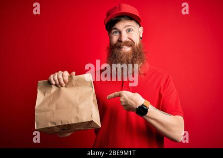 Redhead uomo irlandese consegna con barba tenere take away sacchetto di carta su sfondo rosso molto felice indicando con mano e dito Foto Stock