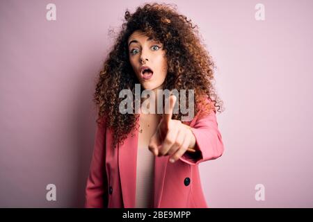 Giovane bella donna d'affari con capelli ricci e piercing indossare elegante giacca puntamento dispiaciuto e frustrato alla fotocamera, arrabbiato e furioso Foto Stock
