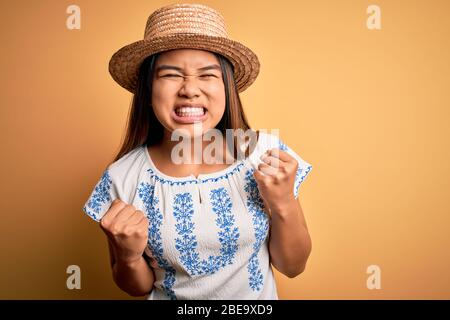 Giovane bella ragazza asiatica che indossa casual t-shirt e cappello in piedi su sfondo giallo celebrazione sorpreso e stupito per il successo con le braccia sollevare Foto Stock