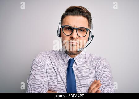Giovane operatore di call center uomo d'affari con occhi blu che indossano occhiali e cuffie skettic e nervoso, disapprovando l'espressione sul viso con a croce Foto Stock