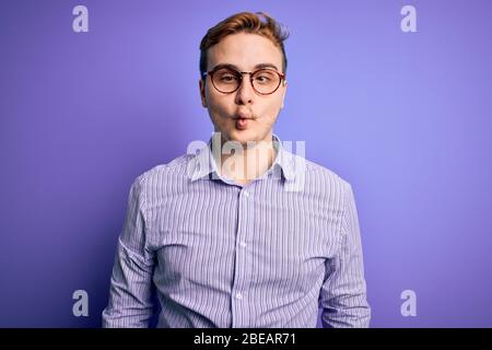 Giovane uomo bello rosso che indossa camicia casual e occhiali su sfondo viola rendendo il pesce faccia con labbra, gesto pazzo e comico. Divertimento Foto Stock