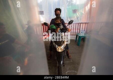 Giacarta, DKI Jakarta, IDN. 13 Aprile 2020. Persone che indossano maschere protettive che attraversano una motocicletta attraverso una camera di disinfezione presso un insediamento densamente popolato a nord di Giacarta, Indonesia. Alcuni residenti a Giacarta hanno iniziato a combattere seriamente il coronavirus (COVID-19) facendo un blocco e anche fornire vari modi, come la camera di disinfezione negli insediamenti stradali di accesso. Credit: Muhmmad Zaenuddin/ZUMA Wire/Alamy Live News Foto Stock