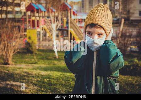 Un ragazzo in una maschera protettiva si trova vicino ad un parco giochi in strada durante la pandemia di coronavirus e Covid - 19 Foto Stock