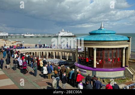 Il banchere pubblico sul lungomare di Eastbourne che tiene un concerto della Brass Band durante un pomeriggio primaverile con il molo in lontananza, East Sussex Inghilterra UK Foto Stock