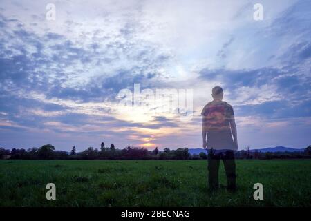 Un escursionista maschile trasparente con zaino con retro a macchina fotografica guardando il tramonto in campagna. Foto Stock