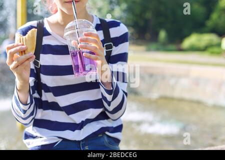 Cibo di strada, ragazza adolescente che mangia sandwich e bere da vetro con paglia. Foto Stock