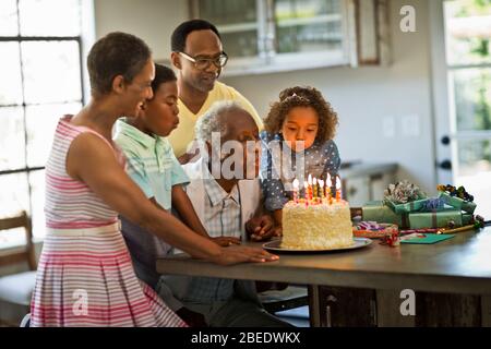 Uomo anziano che soffia fuori le candele sulla sua torta di compleanno con i suoi nipoti ad un raduno della famiglia Foto Stock
