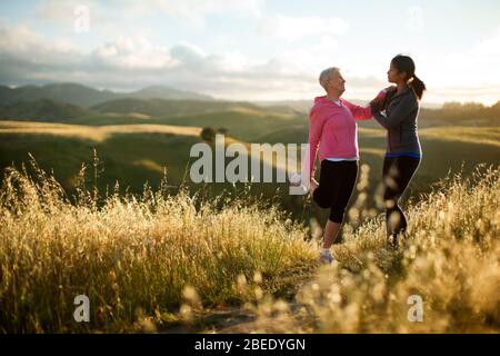 Una giovane donna che aiuta una donna anziana a allungarsi mentre si esercita in terreno collinare Foto Stock