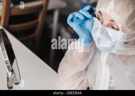 Ragazza in tuta trasparente bianca protettiva, guanti di gomma blu, maschera medica si siede a casa del tavolo e dipinge ciglia con mascara allo specchio durante Foto Stock