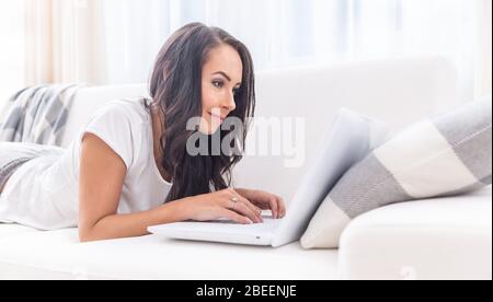 Bella giovane femmina sorridente in una t-shirt bianca e pantaloni sudorati sdraiati su un divano, scrivendo su un laptop appoggiato sul cuscino. Foto Stock