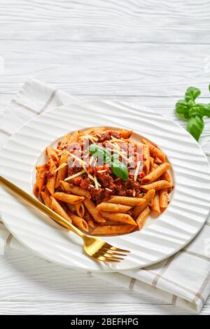 primo piano di pasta bolognese di carne di grano integrale rigata ricoperta di formagio grattugiato e basilico su un piatto bianco, su un tavolo di legno, vista verticale da Foto Stock