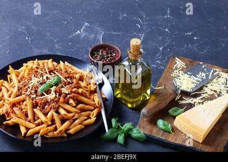 pasta di grano integrale penne bolognese condite con parmigiano grattugiato e basilico su un piatto nero su un tavolo di cemento con ingredienti, vista orizzontale da un Foto Stock