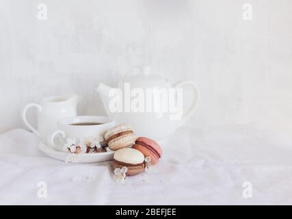 Gustosi macaroni francesi e un set da tè con rami fioriti su sfondo bianco. Colori pastello. Sfondo di primavera. Foto Stock