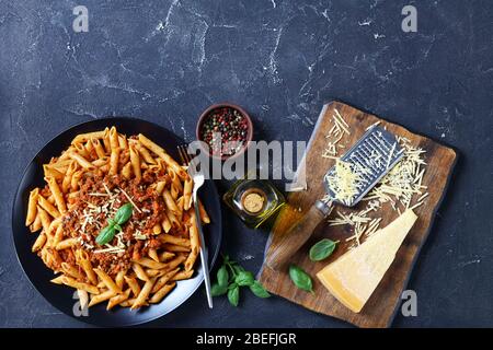 pasta di grano integrale penne bolognese condite con parmigiano grattugiato e basilico su un piatto nero su un tavolo di cemento con ingredienti, vista orizzontale da un Foto Stock