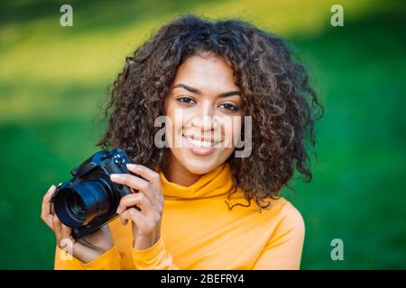 Giovane donna africana graziosa in giallo scatta le immagini con la macchina fotografica di DSLR su sfondo verde. Ragazza sorridente divertirsi come fotografo. Foto Stock