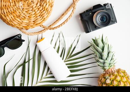 Accessori da viaggio piatti su sfondo bianco con foglia di palma tropicale. Fotocamera con vista dall'alto, occhiali da sole, borsa in rattan, ananas, lozione solare. Somma Foto Stock