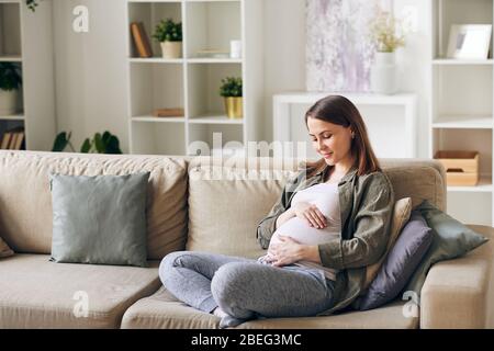 Calma attraente giovane donna incinta seduta con gambe incrociate sul divano e abbracciando il suo ventre Foto Stock
