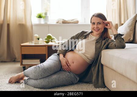 Ritratto di giovane donna incinta sorridente seduta a terra e appoggiata sul divano mentre riposava in congedo di maternità a casa Foto Stock