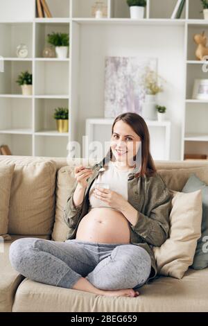 Ritratto di donna giovane incinta positiva seduta con gambe incrociate sul divano e mangiare yogurt naturale a casa Foto Stock