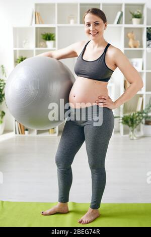 Donna in gravidanza giovane e positiva con leggings grigi e reggiseno sportivo in piedi con flipper su tappetino yoga a casa Foto Stock