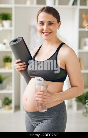 Ritratto di giovane donna allegra incinta in grigio abbigliamento sportivo in piedi con rolled yoga mat e bottiglia fitness, si prende cura della sua salute Foto Stock