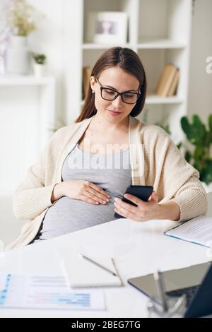 Giovane donna d'affari incinta in occhiali seduti alla scrivania e controllo messaggero telefonico in ufficio Foto Stock