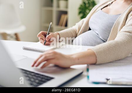 Primo piano di donne incinte irriconoscibili seduti al tavolo e utilizzando il laptop mentre scrivono le attività in blocco note Foto Stock