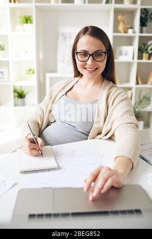 Sorridente, graziosa donna incinta negli occhiali seduti alla scrivania con carta da grafico e appunti mentre si analizzano le informazioni sul laptop Foto Stock