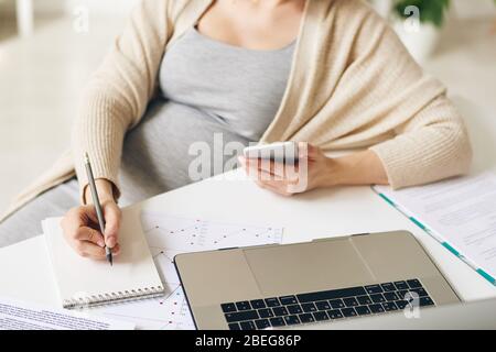Primo piano di una donna incinta irriconoscibile con smartphone seduto al tavolo e prendendo appunti sulle attività durante la visualizzazione dei dati sul laptop Foto Stock
