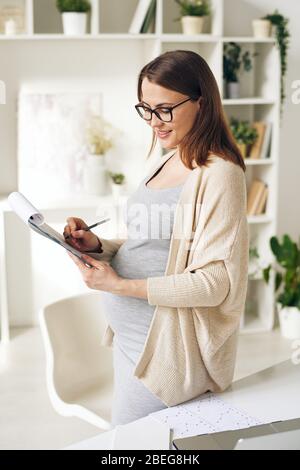 Sorridente donna incinta attraente in cardigan e occhiali in piedi alla scrivania e prendere appunti in carte mentre la analizzano in ufficio domestico Foto Stock