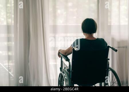 Premuto triste donna in sedia a rotelle usurati guardando fuori dalla finestra e pensare, lonely disabile adulto persona caucasici in ambienti interni Foto Stock